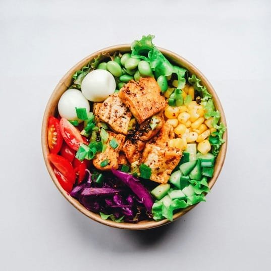 A colorful salad bowl with grilled chicken, tomatoes, purple cabbage, lettuce, cucumbers, edamame, corn, and boiled eggs is a perfect choice to avoid mindset mistakes that often lead to binging.