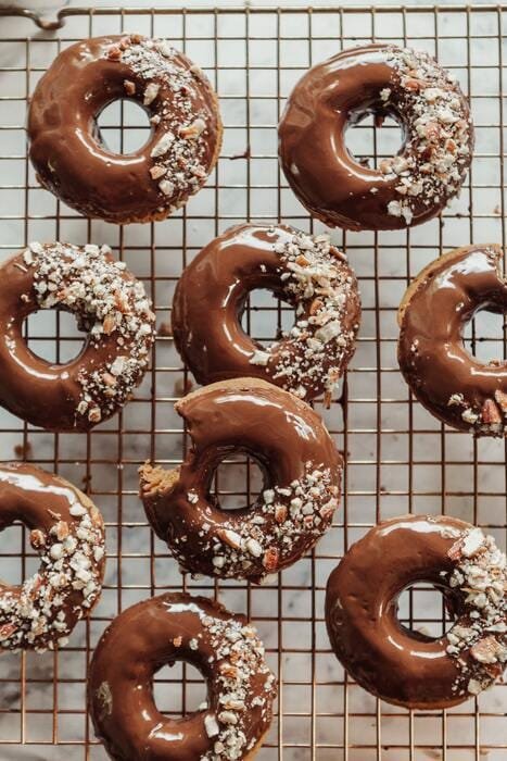 Nine chocolate-glazed donuts, some topped with crushed nuts, are arranged on a cooling rack—a tempting treat that could be enjoyed as part of your Food Peace Reset Programme; one donut has a bite taken out of it.